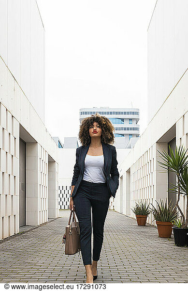 Gut gekleidete Geschäftsfrau mit Handtasche auf dem Fußweg inmitten eines modernen Bürogebäudes