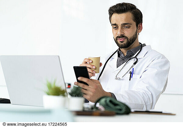 Gutaussehender männlicher Angestellter im Gesundheitswesen  der Kaffee trinkt und ein Mobiltelefon benutzt