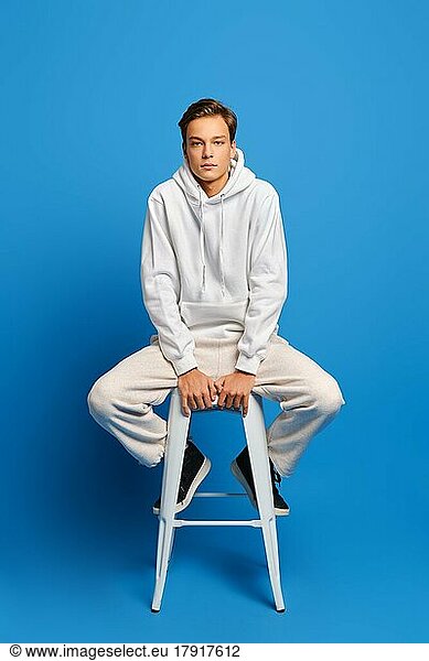 gutaussehender Mann in weißem Sweatshirt und Jogginghose sitzt auf einem weißen Stuhl vor blauem Studiohintergrund