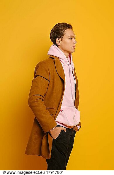 gutaussehender  stilvoller Mann in braunem Trenchcoat und rosa Sweatshirt mit Kapuze im Profil vor gelbem Studiohintergrund stehend