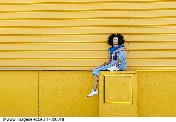 Hübsche Frau sitzt auf einem Podest vor einer gelben Wand und lächelt