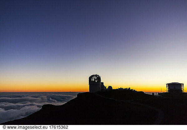 Haleakala Observatory buildings above the clouds along the top of a silhouetted landscape with a golden sunset; Maui Hawaii United States of America