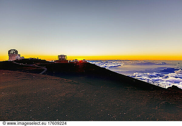 Haleakala Observatory buildings above the clouds with a golden sunset and a view of the ocean and coastline below; Maui  Hawaii  United States of America