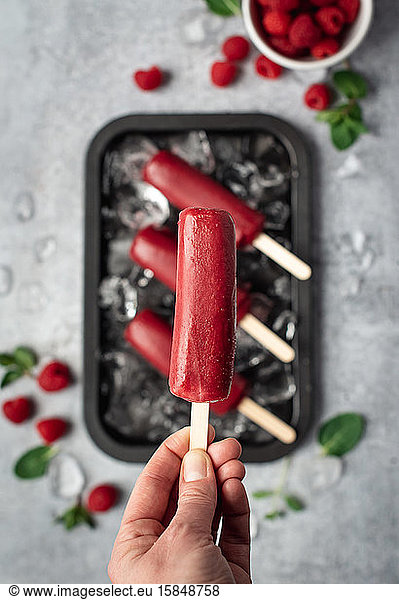 Hand holding raspberry popsicle above a tray of frozen treats.