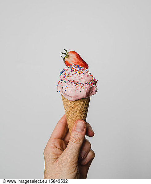 Hand holding strawberry ice cream cone against white background.