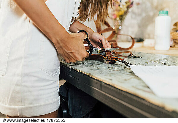 Hand of craftsperson cutting leather belt at workplace
