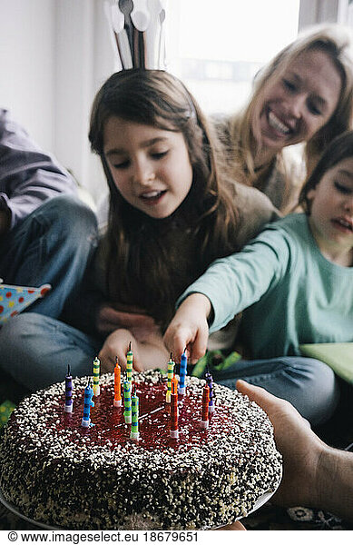 Hand of man holding birthday cake with candles against family at home