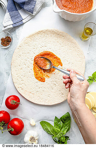 Hand of mature woman spreading tomato sauce with spoon on pizza dough