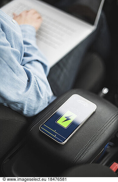 Hand of mature woman using laptop with electric car charging app on smart phone screen