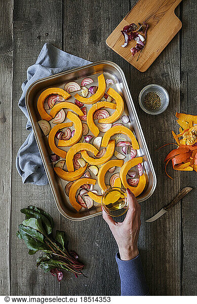 Hand of woman sprinkling olive oil on sliced onions  pumpkins and common beets on baking sheet