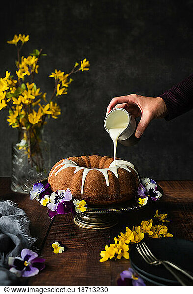 Hand pouring icing on bundt cake decorated with flowers.