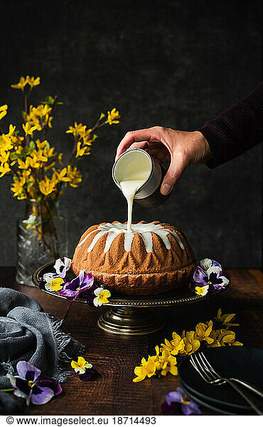 Hand pouring icing on bundt cake decorated with flowers.