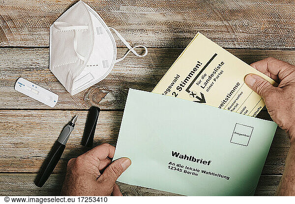 Hand putting ballot for federal elections into envelope on table with protective mask and corona rapid test