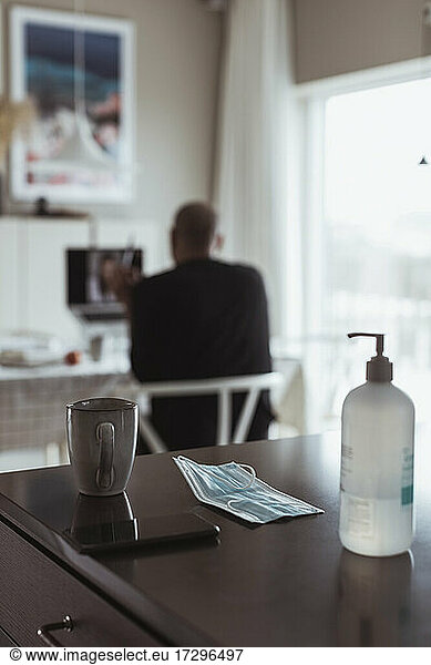 Hand sanitizer and mask on table while male professional in background at home during pandemic