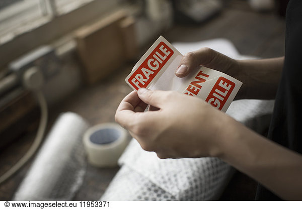 Hands holding a red Fragile sticker to stick it on a brown paper package on a work bench.