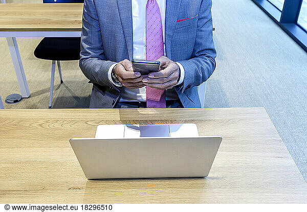 Hands of businessman using mobile phone sitting with laptop at desk in office