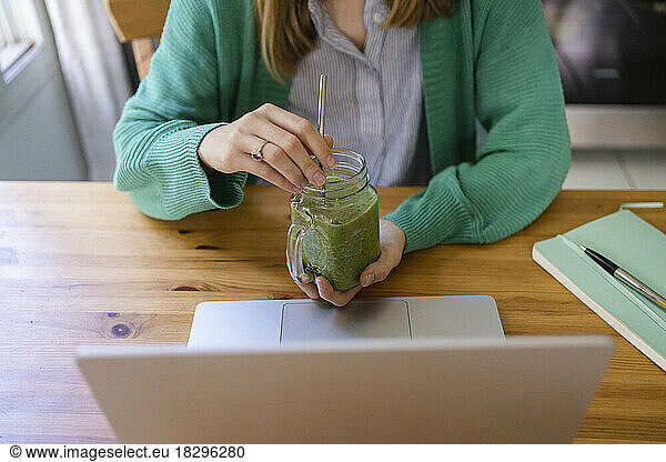 Hands of freelancer holding green smoothie with laptop on table