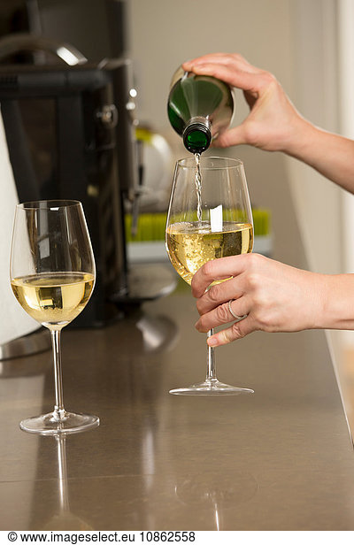 Hands of woman pouring glasses of white wine at kitchen counter