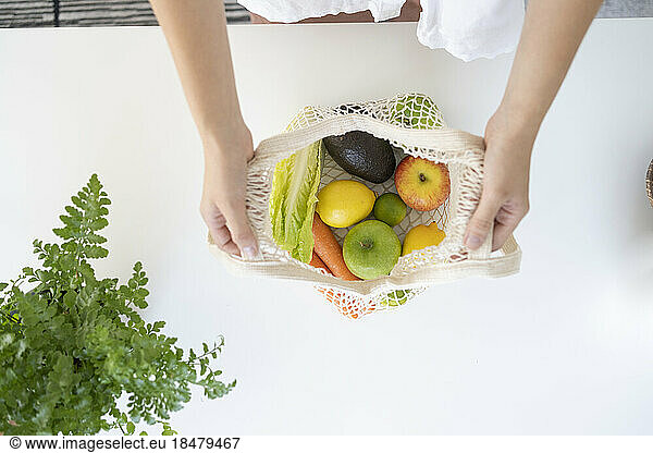 Hands of woman with fruits in mesh bag on table at home