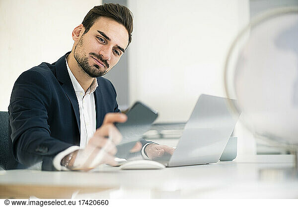Handsome male professional checking smart phone while sitting with laptop at desk in office