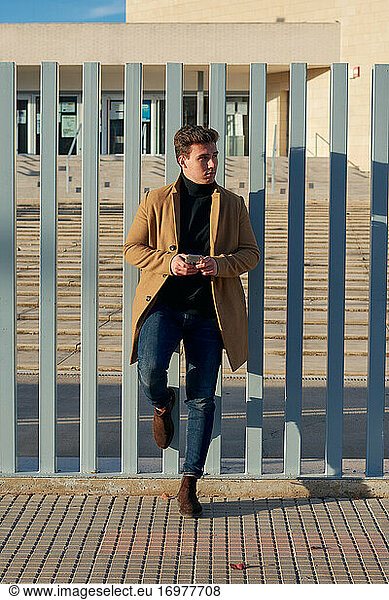 Handsome man dressed in a coat holds his mobile leaning on a fence