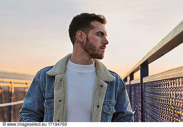 Handsome man in a jacket poses on a bridge at sunset