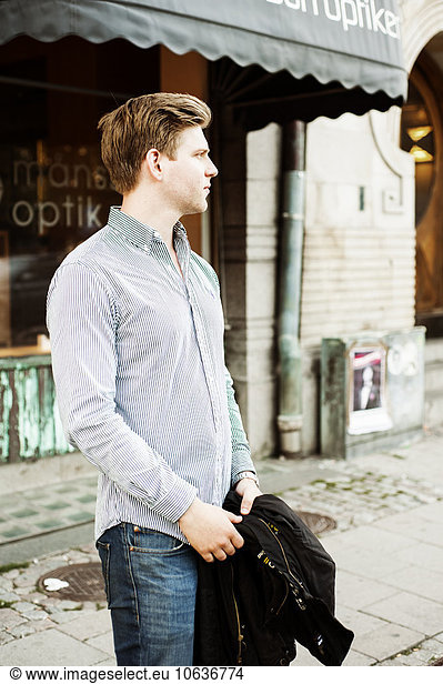 Handsome man looking away while holding jacket in city