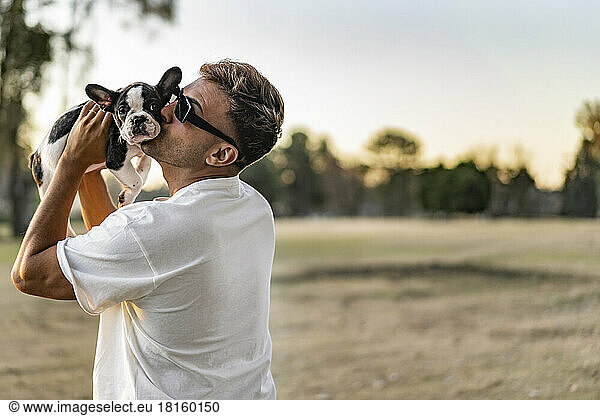 Handsome young man holding up and kissing a french bulldog.