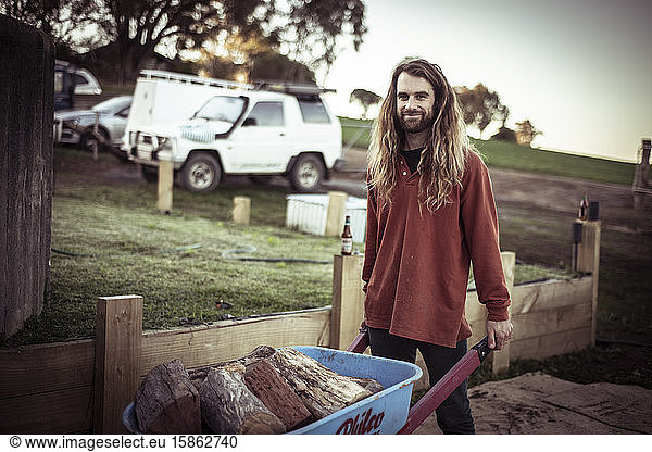 handsome young man with beard and long hair pushes firewood on farm