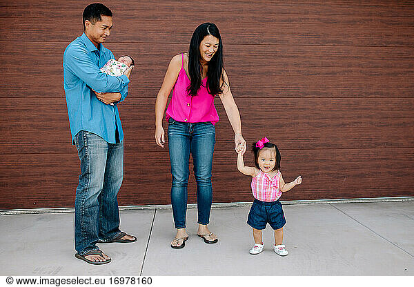 Happy asian mom holds hand of toddler while dad carries newborn baby