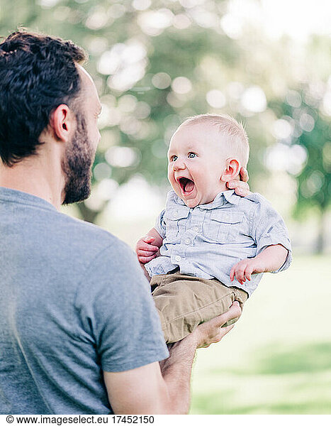 Happy Baby Boy Laughing as Dad Lifts Him in Air at Park in Summertime