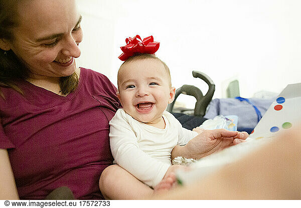 Happy Baby Sits in Mother's Lap Smiling with Red Bow on Head