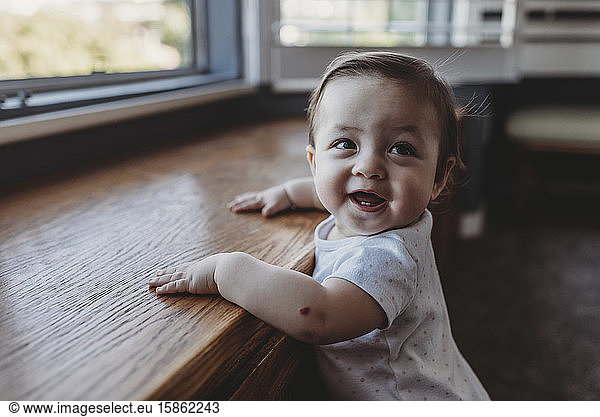 Happy baby with first teeth holding herself upright near window