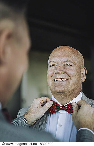 Happy bald gay man looking at partner adjusting bow tie before wedding