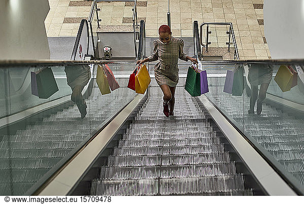 Happy black woman on a funny haircut holding colorful shopping bags walking on the escalators