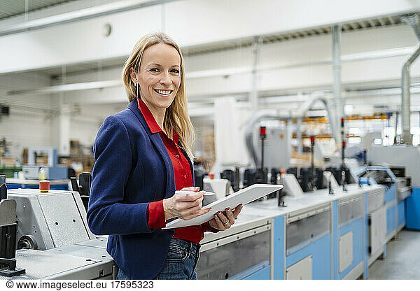 Happy blond businesswoman with tablet PC standing by machinery in industry
