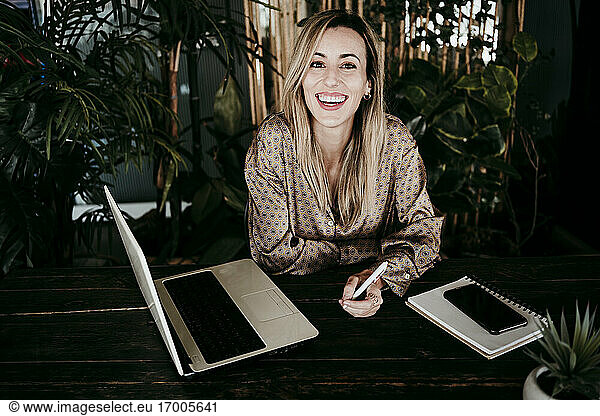Happy blond female entrepreneur sitting with laptop at desk in office