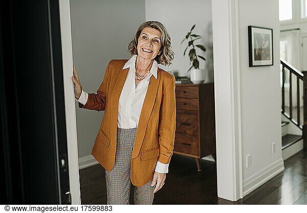Happy blond senior businesswoman standing at home looking through door