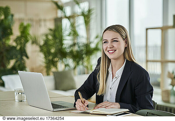 Happy blond teenage intern looking away while sitting with diary at desk in office