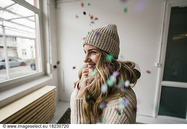 Happy blond woman wearing knit hat standing amidst falling confetti at home