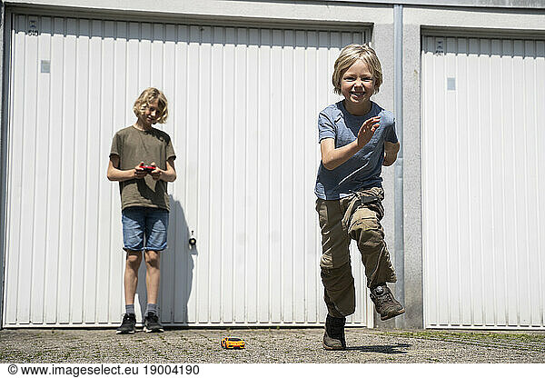 Happy boy dancing and brother playing with remote controlled car in yard outside garage