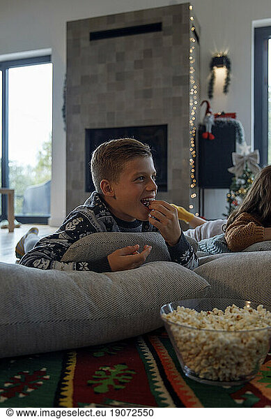 Happy boy eating popcorn by sister and watching TV at home