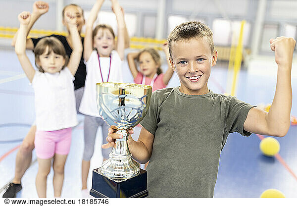 Happy boy flexing muscle holding trophy with friends at school sports court