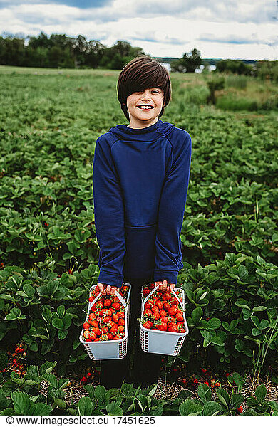 Happy boy holding full baskets of ripe strawberries in a field.