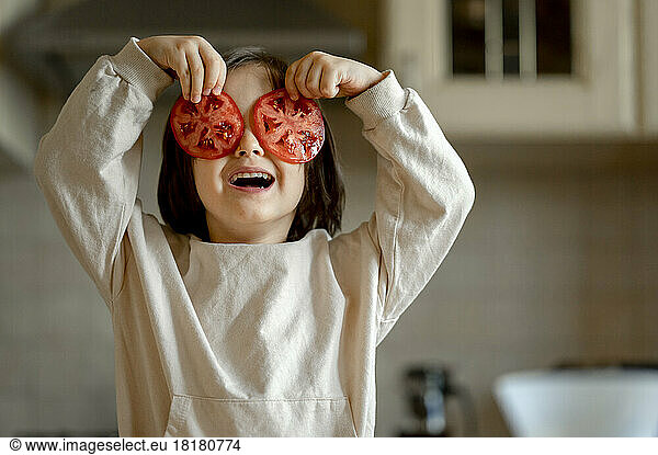 Happy boy holding tomato slices over eyes in kitchen at home