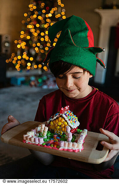 Happy boy in elf hat looking at gingerbread house at Christmastime.