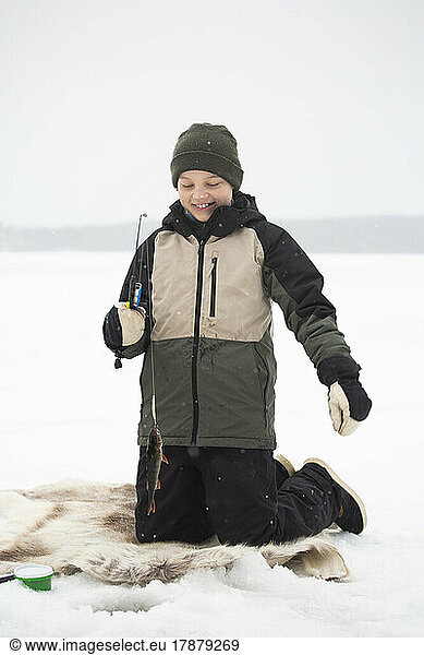 Happy boy in warm clothing kneeling while fishing on frozen lake
