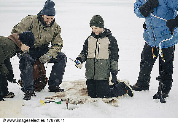 Happy boy kneeling by men and friend while fishing at frozen lake during winter