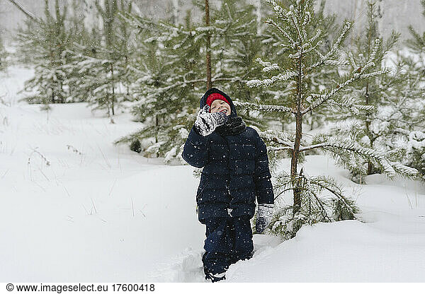 Happy boy wearing warm clothing standing in snow forest by tree