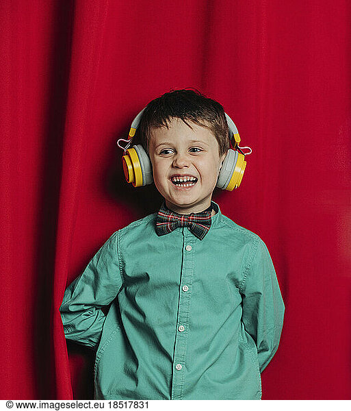 Happy boy wearing wireless headphones standing in front of red curtain